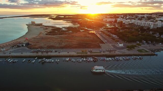 Dramatic aerial view of a ferry boat navigating a channel in the picturesque town of fuseta at sunset, showcasing the stunning algarve coastline and its beautiful golden light over the landscape