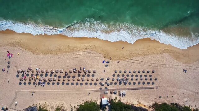 Beautiful aerial top down view of a sandy coastline with rows of straw umbrellas and people relaxing, as turquoise ocean waves gently wash ashore during a sunny summer day