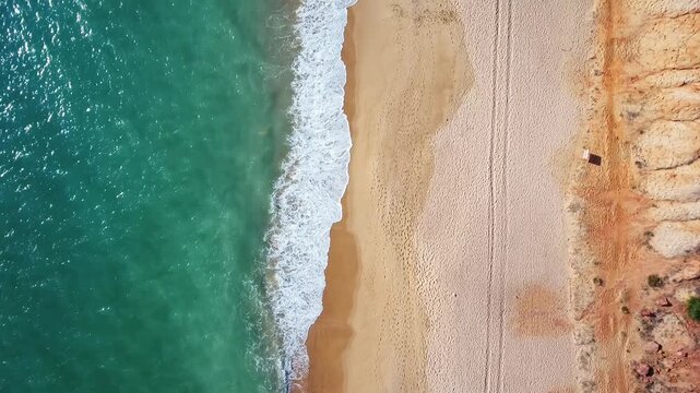 Aerial top down view of a deserted coastline with turquoise ocean waves gently rolling onto a sandy beach next to ochre cliffs, showcasing the serene and pristine beauty of nature