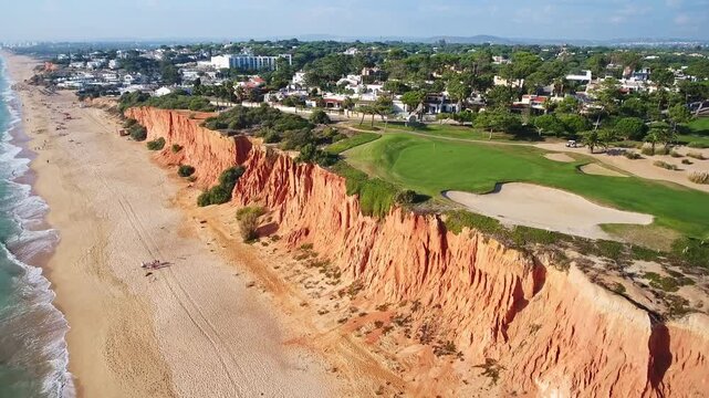Dramatic aerial perspective flying over the beautiful coastline of the algarve region in portugal, featuring stunning red cliffs, a sandy beach, turquoise ocean water, and a lush green golf course