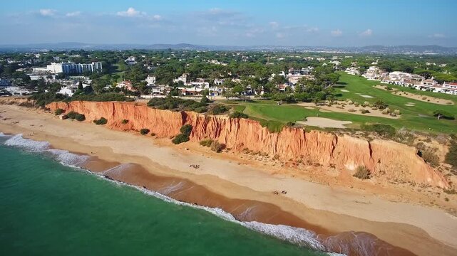 Dramatic aerial footage showing the beautiful red cliffs of garrao beach bordering the green vale do lobo golf course, a popular tourist destination in the algarve region of southern portugal