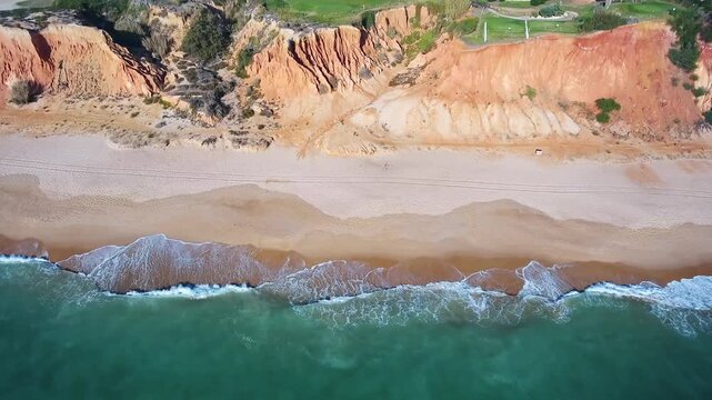 Amazing aerial top down view of turquoise ocean waves gently crashing on a deserted sandy beach next to stunning red and orange cliffs, revealing the natural beauty of the coastline