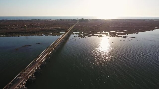 Aerial view of the long wooden bridge in quinta do lago, algarve, stretching over the tranquil ria formosa lagoon towards the beach, with the afternoon sun reflecting on the water surface