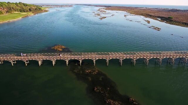 Aerial view revealing the scenic quinta do lago wooden bridge crossing the ria formosa lagoon, with people walking towards the beach and a golf course visible on the shore in algarve, portugal