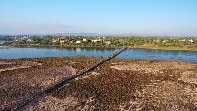Beautiful aerial footage showing the iconic wooden bridge spanning the ria formosa lagoon, connecting the quinta do lago resort area to the beach in the algarve region of southern portugal