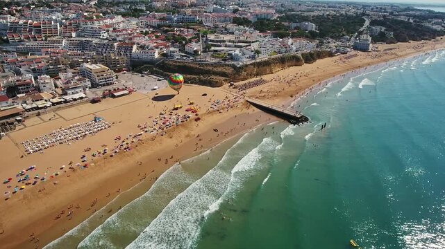 Aerial panorama of albufeira with a colorful hot air balloon flying over the crowded golden sand of fisherman's beach, showing tourists enjoying the turquoise waves of the atlantic ocean