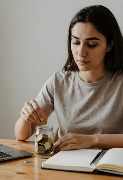 Personal finance and savings strategy, young woman putting coins into a glass jar for budget planning and future investment goals at a home office desk