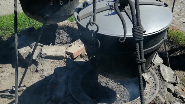 Traditional Cast Iron Cauldron with Lid Hanging on Metal Tripod for Outdoor Cooking Over Fire at Rustic Campsite