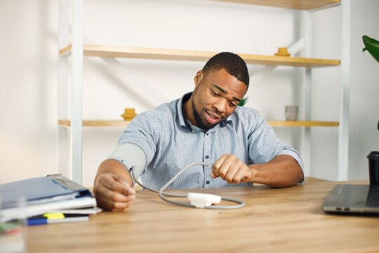 Black male entrepreneur sitting in office near laptop and using tonometr