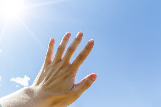 A human hand reaches out towards the bright sun against a clear blue sky on a sunny day.