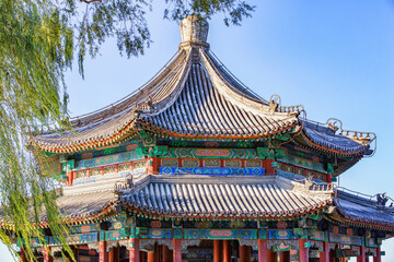 Fototapeta premium A two-tiered traditional Chinese pavilion with ornate painted decorations and curved tile roofs, set outdoors under a clear blue sky with willow branches nearby.