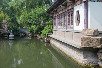 A traditional Chinese pavilion stands by a calm pond, with a small stone bridge and lush greenery in a classic garden setting. © tai