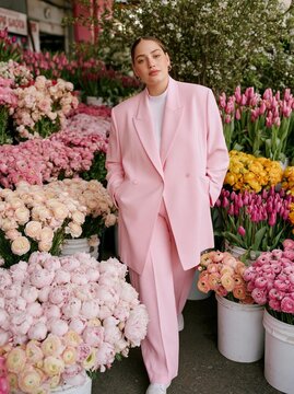 Young woman standing in front of a large display of colorful flowers. she is wearing a bright pink suit with a white turtleneck and matching trousers.