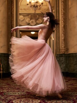 Young ballerina in a pink tutu, gracefully dancing in a grand room with ornate decorations and a chandelier.