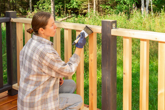 Woman working on a DIY home improvement project outdoors. Applying protective wood care oil to a terrace railing with a paintbrush. Exterior house renovation and deck maintenance concept.