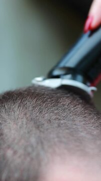 Close-up of a person with red manicure using a hair clipper to buzz-cut short brown hair. Hairdressing, grooming, and barber shop concept