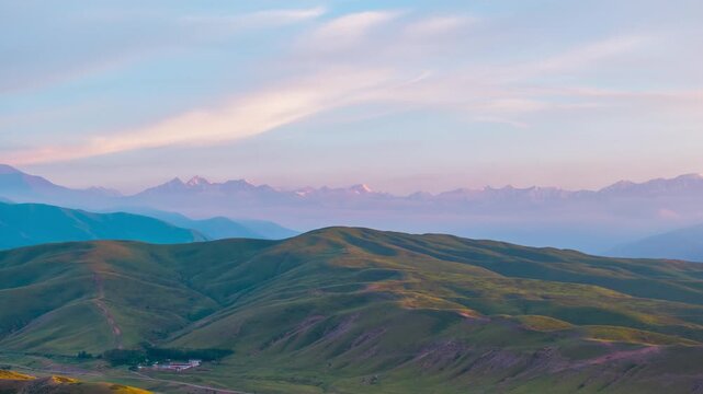 Drone hyperlapse flying forward over green hills and valley settlement toward distant snow peaks under soft dawn sky in Kyrgyzstan.