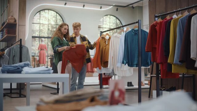 Clothing Store Shopping: Young Caucasian Woman Searches a Rail of Colorful Apparel Before Her Boyfriend Joins Her, Sharing Opinions and Laughing Together During a Casual Fashion Shopping Trip.