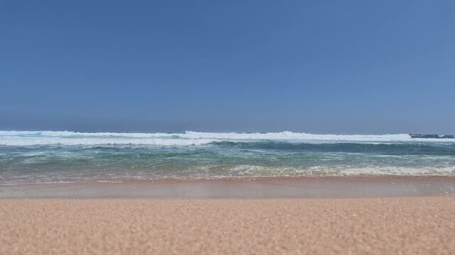Low level perspective capturing crystal clear sea foam washing over fine wet sand with rhythmic ocean waves breaking under a clear blue sky, ideal for summer holiday promos.
