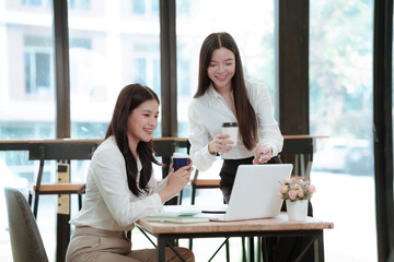 Obraz premium Two young Asian businesswomen celebrating a successful project with a laptop, showing teamwork, achievement, motivation, and business success in a modern office.
