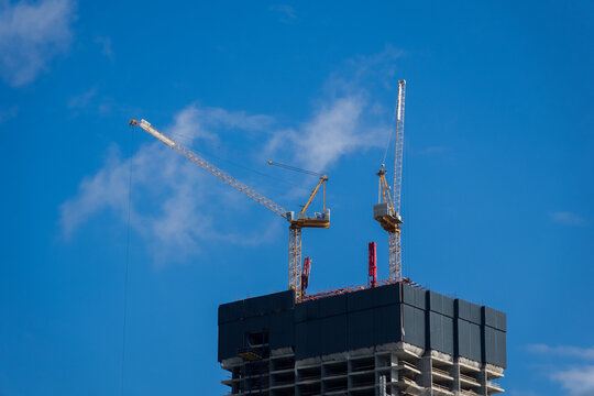 Construction cranes work on a tall building under a blue sky with some clouds in the background during the day