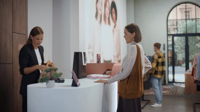 Shopping at Clothing Store, Female Shopper Chooses an Item From a Rack and Carries It to the Cash Desk, Friendly Professional Sales Associate Folds the Garment and Slips It Into a Paper Bag.
