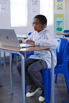 African American youth male sitting at school desk typing on laptop in grey hoodie, blue chair