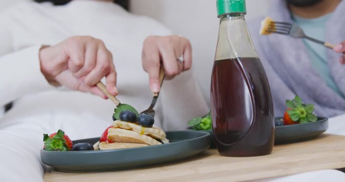 Two adults are pressing fork and cutting blueberry pancakes on wooden lap tray to share breakfast