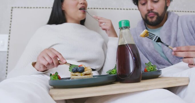 African American couple sharing breakfast in bed, tray on laps, cutting pancakes by green-cap syrup