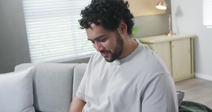 Adult male reading messages and scrolling smartphone while sitting on light-gray sofa, smiling
