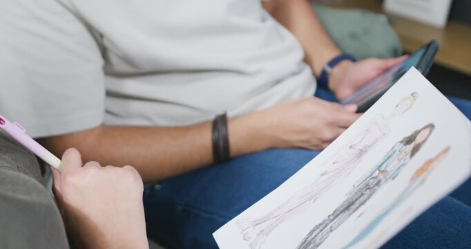 After pen lifting, two designers are pointing at fashion sketches on sofa and comparing with tablet
