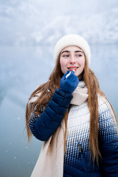 Woman using moisturizing lip balm outdoors in winter, demonstrating cold-weather lip care against a scenic snowy mountain lake background.