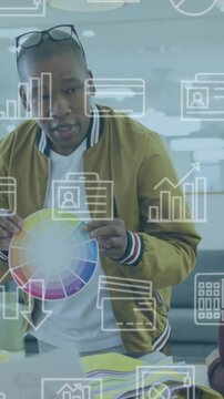 Animation of multiple icons over diverse man showing color swatch to coworkers in meeting at office