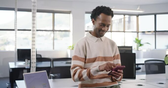 Man in beige sweater leaning at open office desk, tapping purple phone, reading message by laptop