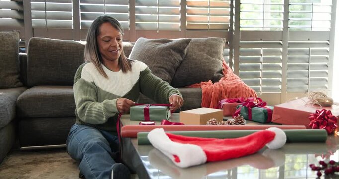 Mature African American woman sitting at home wrapping gifts for holiday with ribbon, copy space