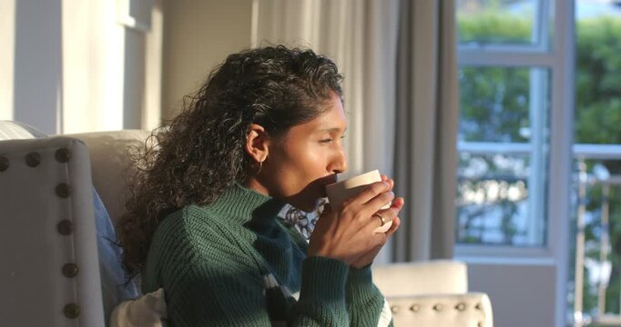 Sunlight drawing African American woman by window, holding mug, lifting to lips, sipping and gazing