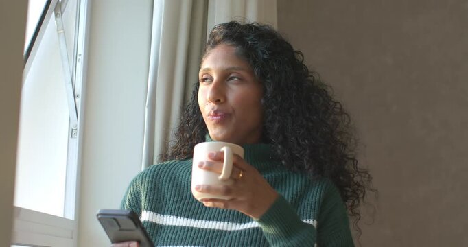 African American woman in green sweater at home sipping cream mug gazing out window scrolling phone