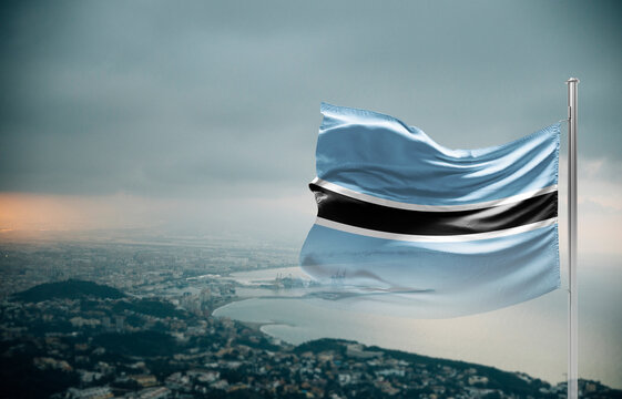 Botswana flag waving over a coastal city under dramatic cloudy sky, realistic fabric motion, aerial seaside landscape background. Botswana national flag for independence day.