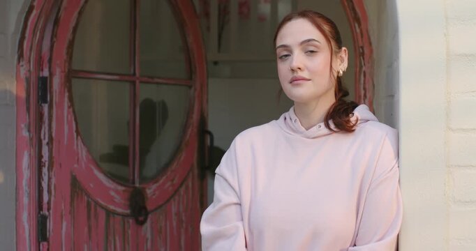 Woman returning gaze to camera, smiling, tucking hair in pink hoodie by red door, copy space