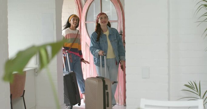 Diverse female friends entering pink arched doorway, pulling beige, black suitcases