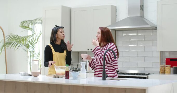 Diverse women right tasting small bowl sparking left in yellow whisking batter at kitchen island