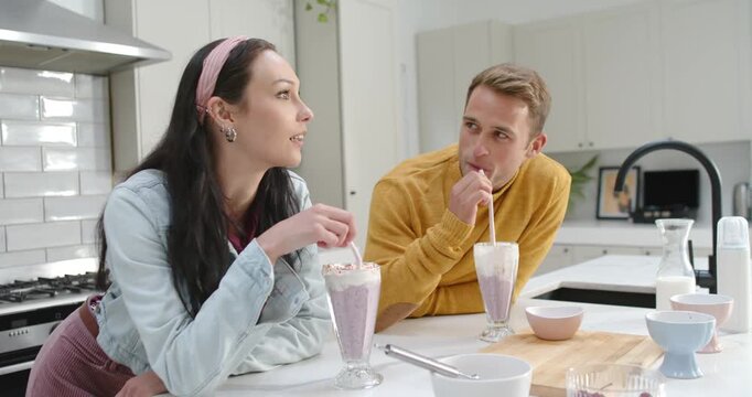Couple leaning on kitchen island with shakes when woman sipping, sharing playful talk and laughing