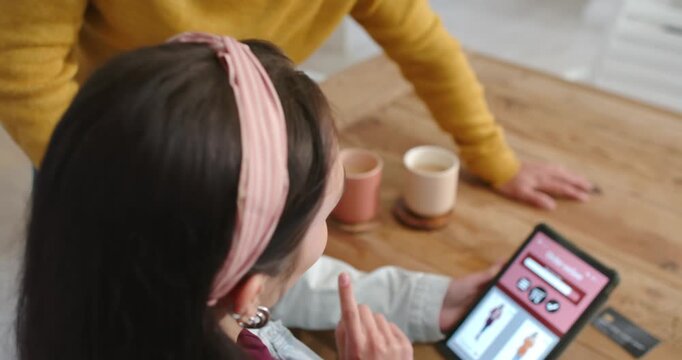 Couple at home dining table, man leaning in sparking woman tapping shopping tablet with mugs, card