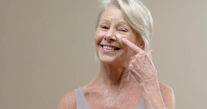 Senior female applying finger cream under right eye after dab appearing, showing skincare in studio