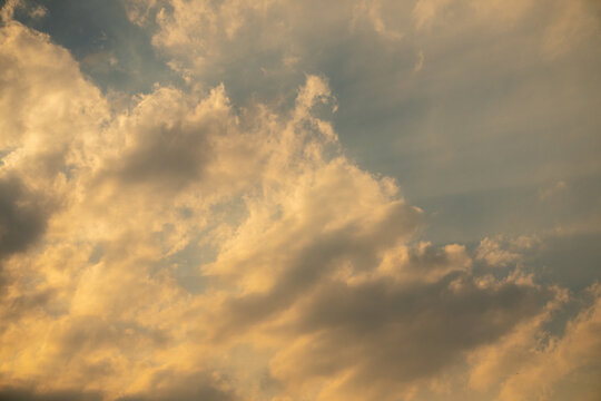 A cloudy sky illuminated by the warm, golden and orange glow of sunset or sunrise. Dense, billowing cloud formations featuring soft shadows and luminous highlights.