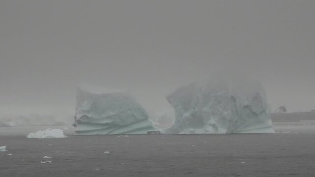Massive iceberg shrouded in fog and low clouds on the Antarctic Peninsula, with a snow-covered coastline fading into the mist