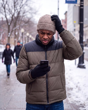A man in a beige knit beanie and two-tone puffer jacket looks concerned at his phone on a snow-covered city street
