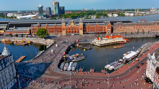 Aerial drone view of Amsterdam Centraal station with crowds of pedestrians and cyclists on Stationsplein, canal cruise boats on the Open Havenfront and the IJ river, Netherlands, in summer.