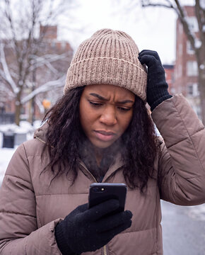 A concerned woman wearing a knit beanie and puffer jacket looks puzzled at her smartphone in a snowy urban setting