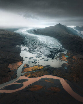 Massive glacier flows through a dark mountain valley into braided meltwater streams, creating a dramatic aerial landscape ideal for climate, geology, travel and environmental content.